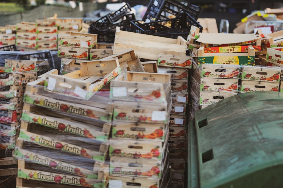 A large collection of wooden fruit boxes stacked in an outdoor area, with some black plastic crates piled behind them. The cardboard boxes are decorated with colorful images and text indicating they are used for fresh fruits, featuring illustrations of apples, tomatoes, and other produce. The wooden boxes have a light, natural finish with some showing visible grain and slight wear. They are arranged in uneven stacks on a pavement surface, with some boxes leaning against each other. Surrounding the boxes are a few black plastic crates and a portion of a green waste bin or container in the foreground. The environment appears to be a public or commercial space, possibly a market or delivery yard, where waste clearance operations by companies like Waste Clearance Hackney may be involved in removing packaging debris. The lighting is natural, likely outdoors during daytime, highlighting the textures of the wood and cardboard materials, supporting efficient waste management or alternative disposal methods related to rubbish removal services.