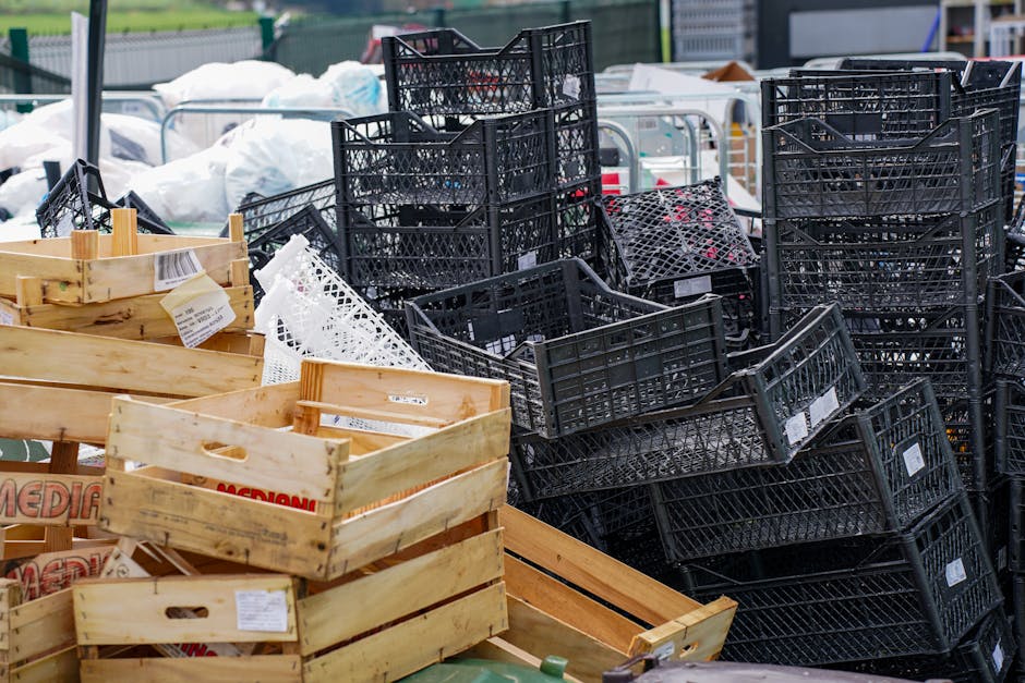 A large collection of wooden fruit boxes stacked in an outdoor area, with some black plastic crates piled behind them. The cardboard boxes are decorated with colorful images and text indicating they are used for fresh fruits, featuring illustrations of apples, tomatoes, and other produce. The wooden boxes have a light, natural finish with some showing visible grain and slight wear. They are arranged in uneven stacks on a pavement surface, with some boxes leaning against each other. Surrounding the boxes are a few black plastic crates and a portion of a green waste bin or container in the foreground. The environment appears to be a public or commercial space, possibly a market or delivery yard, where waste clearance operations by companies like Waste Clearance Hackney may be involved in removing packaging debris. The lighting is natural, likely outdoors during daytime, highlighting the textures of the wood and cardboard materials, supporting efficient waste management or alternative disposal methods related to rubbish removal services.