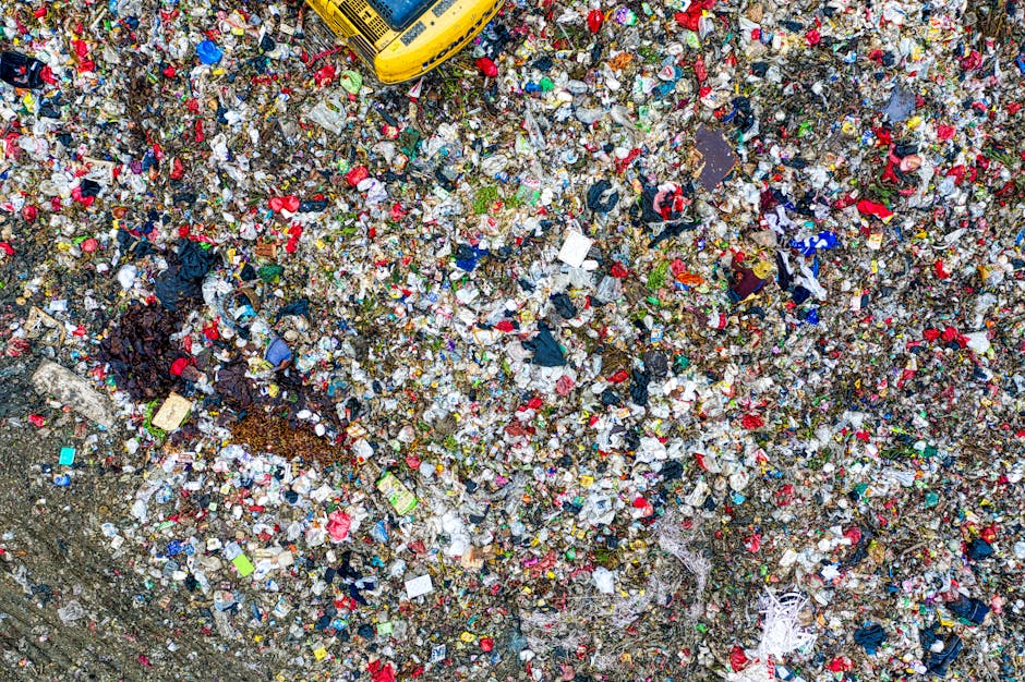 An aerial view of a large, densely packed waste site filled with mixed household and commercial rubbish. The ground is covered with a thick layer of assorted waste materials, including plastics, paper, fabric, cardboard, and food packaging, creating a colorful and chaotic surface. In the upper part of the image, a yellow waste collection vehicle is partially visible, indicating ongoing rubbish removal or transfer processes. The waste appears to be scattered across a dirt or gravel surface, which has small patches of soil visible around the edges. Some items, such as plastic bags and crumpled containers, are piled in certain areas, suggesting recent or ongoing clearing activity. The environment suggests an industrial or disposal area used for large-scale waste management, typical of independent rubbish collection services like Waste Clearance Hackney, who often handle household and commercial debris not processed through local authority waste services. The overall scene highlights the volume and diversity of waste materials encountered during private rubbish clearance operations, especially in urban environments such as Clapton, Hackney.