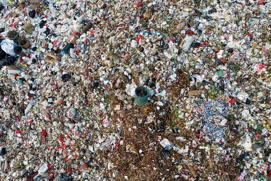 A large dirt and gravel area covered with a dense layer of mixed household and construction waste, including plastic bottles, paper, cardboard, and small discarded items. Among the debris, there are scattered crumpled plastic bags, pieces of torn fabric, and various unidentifiable rubbish materials in different colours such as red, white, and blue. The ground appears uneven with piles of waste creating a rough textured surface, and some areas have compacted soil visible beneath the waste. Partial views of wooden pallets and a small blue plastic bucket can be seen protruding from the rubbish, indicating a site used for waste collection or temporary storage. The environment is outdoors, possibly a waste yard or an unmanaged disposal site, with no visible structures or vegetation nearby. This scene exemplifies the kind of clutter associated with private or independent rubbish clearance services, like those offered by Waste Clearance Hackney, focusing on the removal of accumulated domestic and construction waste to manage incomplete or improper disposal practices.