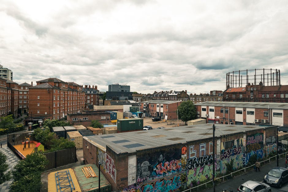 A cityscape view showing a variety of urban buildings, including mid-rise and low-rise brick structures, with some featuring flat metal roofs and others with tiled roofs. In the foreground, there is a large, single-story industrial building with a graffitied brick wall, displaying colorful tags and murals. Adjacent to this building, several small sheds and storage containers are visible, some painted in muted tones with corrugated metal surfaces. A few trees and patches of greenery are interspersed among the structures, adding natural contrast to the predominantly brick and metal environment. On the left side, there is a small outdoor play area with a yellow and red slide, enclosed by a fence, suggesting a mixed-use space within the urban setting. In the background, more residential and commercial buildings extend toward the cloudy sky, with an elevated, mock-industrial structure constructed from metal and situated on the right. The scene is well-lit by natural daylight, highlighting the textures of the building materials, which include weathered bricks, painted surfaces, and metal roofs, conveying an impression of an active urban environment where waste removal services such as those offered by Waste Clearance Hackney are essential for maintaining cleanliness and order in densely populated areas.