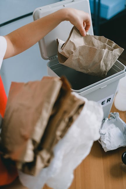 A person is shown disposing of paper waste into a white paper recycling bin that is partially open, situated on a wooden surface indoors, with their arm extended to place a crumpled paper bag inside. In the foreground, there are additional folded and crumpled paper bags held in the person's other hand, emphasizing waste collection. The recycling bin is positioned near a white wall and a black corner furniture piece, with some loose paper waste lying nearby on the surface. The scene depicts typical domestic waste handling, closely related to private rubbish removal services such as those offered by Waste Clearance Hackney, highlighting the importance of proper waste segregation and disposal methods for recyclable materials in urban environments.