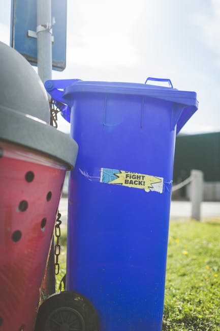 A large, upright blue plastic wheelie bin with a closed lid, situated outdoors on a grassy area. The bin has a sticker with the words 'FIGHT BACK!' in bold, black letters on a yellow background with blue and black lightning bolt graphics. The bin appears clean and is positioned beside other containers, including a red bin with a grey lid that has ventilation holes, and a small black wheel visible at the bottom. In the background, there is a partly cloudy sky, a metal pole with a sign or fixture attached, and a wooden barrier or fence post, indicating an urban or suburban environment. The scene suggests an area used for rubbish or waste collection, consistent with independent or private waste management services such as those provided by Waste Clearance Hackney, supporting alternative waste disposal methods like on-site or non-local authority rubbish removal.