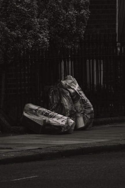A collection of large rubbish bags made from thick, black plastic, tightly packed and leaning against a dark wooden fence on a city pavement. The bags appear to contain household or garden waste, with some showing signs of being filled and sealed, and others slightly bulging from contents. The scene is set outdoors, with a tree visible in the upper left corner and the fence extending across the background. The pavement surface is concrete, and a curb separates the pedestrian area from the road in the foreground. Overhead lighting suggests early morning or late evening conditions, creating a subdued, shadowed atmosphere. This image exemplifies a typical scenario where private waste clearance by companies like Waste Clearance Hackney would be employed for on-site rubbish removal, highlighting the need for professional collection services to handle such waste efficiently and responsibly in urban environments.