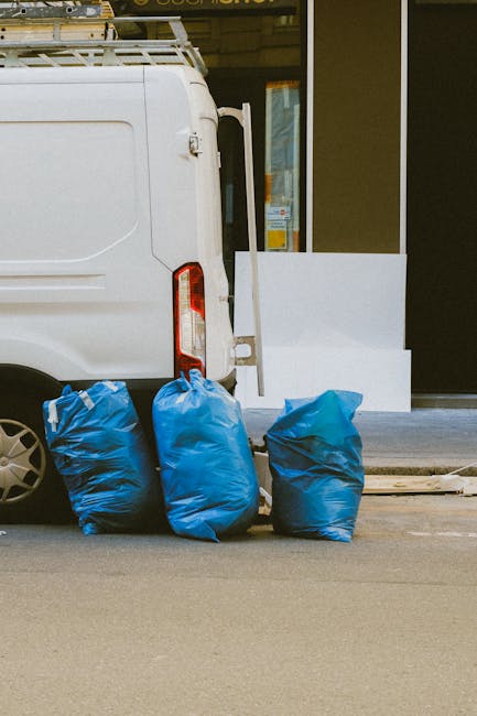 The image shows the rear and side of a white commercial van parked on an urban street, with its back doors closed. Three blue rubbish bags, made of plastic with slightly crinkled textures, are placed on the pavement directly next to the van, leaning against its rear wheel arch. The bags appear to be filled with waste and are of similar size, with some slight variations in shape. In the background, there is a storefront with large glass windows and dark framing, and a white panel or board positioned on the sidewalk, possibly part of a shop front or advertising display, partially visible on the right. The scene is lit with natural daylight, and the setting suggests a typical city environment where independent waste collection, such as that provided by Waste Clearance Hackney, occurs outside businesses opting for private rubbish removal rather than council-led services. The area is clean, and the overall context aligns with alternative waste handling or on-site clearance activities in an urban commercial area.