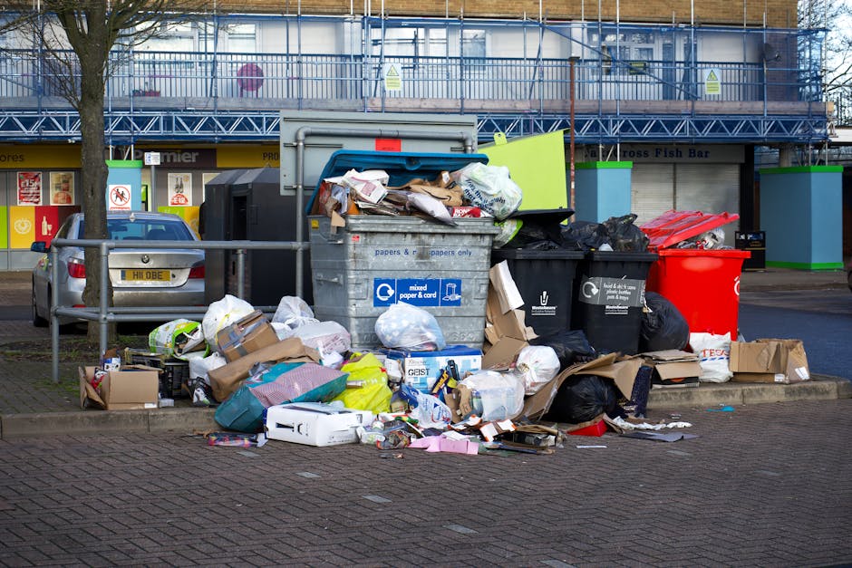 An overflowing collection of mixed waste sits on a paved area with scattered cardboard boxes, plastic bags, and discarded paper among other rubbish. The waste includes crumpled paper, empty packaging, and small household items, with some items spilling onto the adjacent curb and pavement. Behind the rubbish, there is a large metal recycling bin labeled for mixed paper and card, partially open with waste protruding. To the left, a parked silver car is visible behind a low metal railing, and in the background, a commercial building with a blue scaffolding structure covers an upper floor, with shopfront signage including a fish and chip shop. Several black and red waste bags are placed next to the bin, and a few cardboard boxes are flattened or crumpled. The environment is an urban street scene, potentially in Hackney Wick, offering a snapshot of private waste collection practices outside of local authority rubbish disposal, aligning with alternative rubbish removal options. The overall lighting is natural and slightly overcast, highlighting the mess and the surrounding infrastructure.