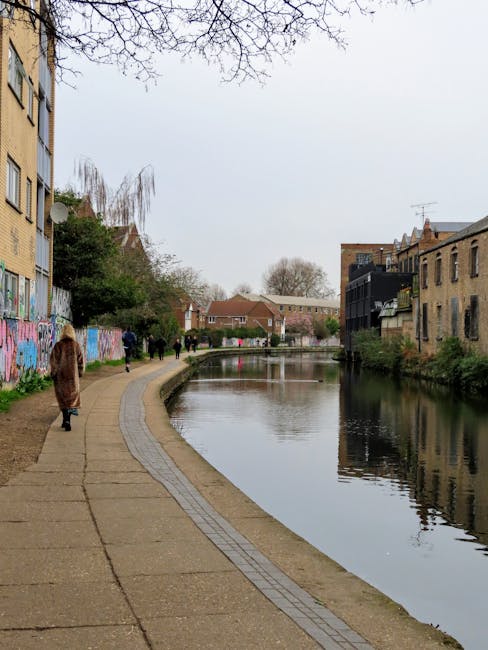 A paved pedestrian walkway runs alongside a narrow canal in an urban setting, with several people walking in the distance. On the left, a tall, yellow brick residential building is visible, with weathered graffiti on its base wall, while green trees and sparse foliage extend over the pathway. The canal is calm, with gently rippled water reflecting the overcast sky and nearby buildings, including a mix of older brick structures and modern black balconies on the right. In the background, trees and low-rise houses can be seen, indicating a mixed residential area. The overall scene suggests a peaceful daytime setting in a city with an accessible riverside or canal-side walk, often utilized for outdoor leisure or alternative waste collection methods associated with local rubbish clearance services, as provided by Waste Clearance Hackney, although no waste is visible in the image. The event of walking and the environment imply an organized public space complementing urban rubbish removal operations related to private waste handling.  